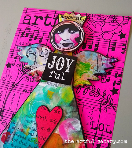 A smiling art angel made out of paper and chipboard wearing a crown and a Joyful tag.